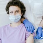 Pielęgniarka w maseczce podaje dorosłej kobiecie szczepionkę w ramię